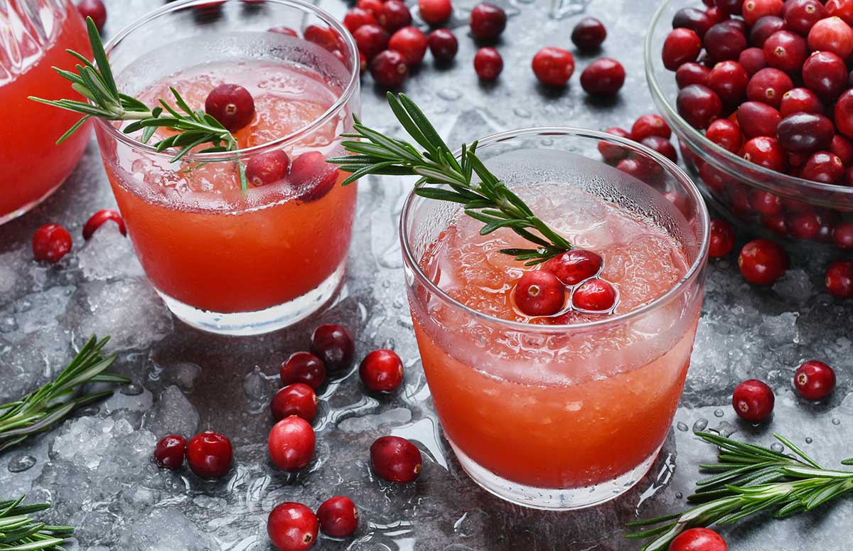 Two glasses of cranberry 国产xxxx视频在线观看 with fresh cranberries and rosemary sprigs, bowl of fresh cranberries, loose cranberries and sprigs on a dark marble countertop.